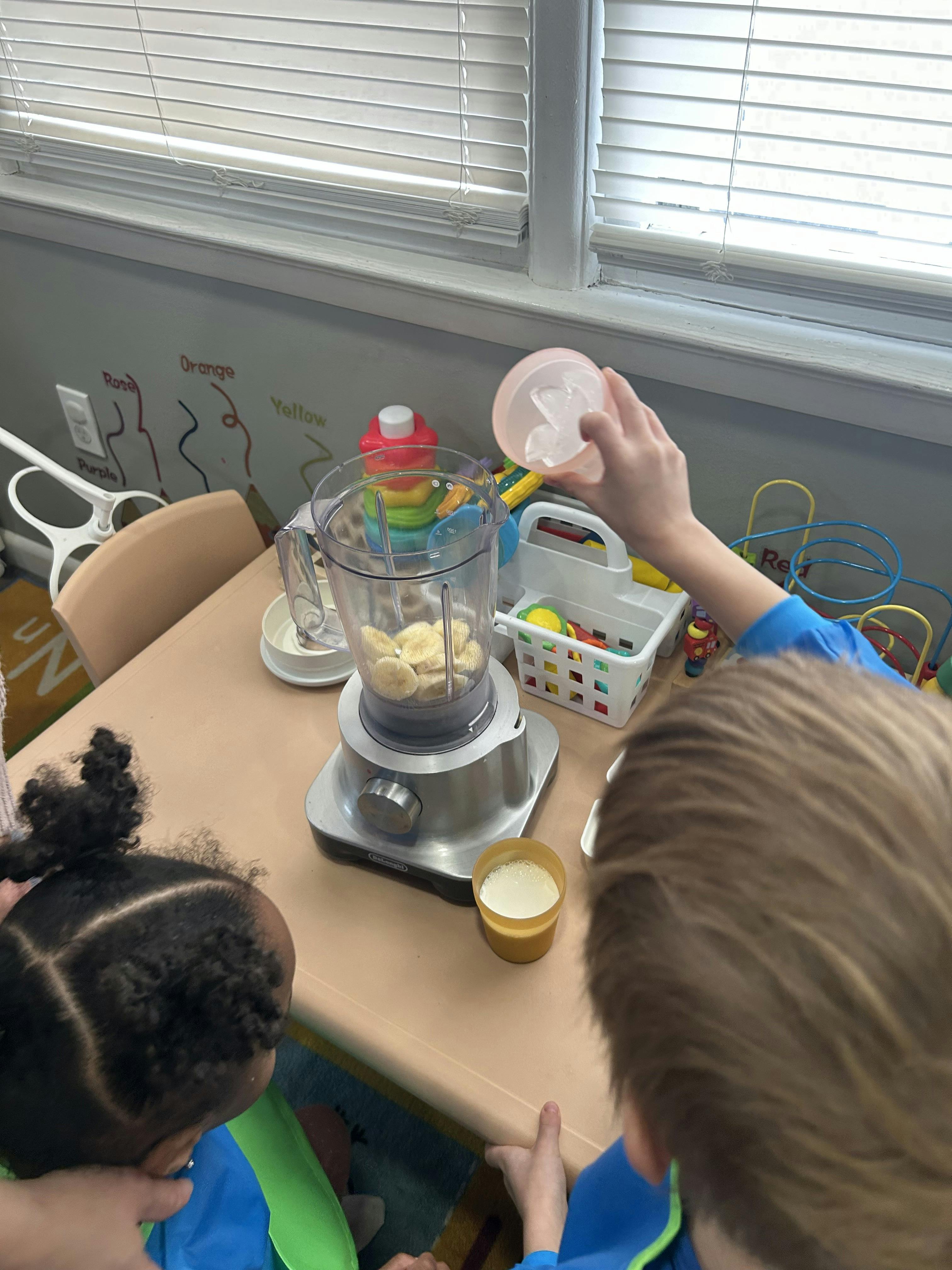 Child adding ice to a blender filled with banana slices as another child watches