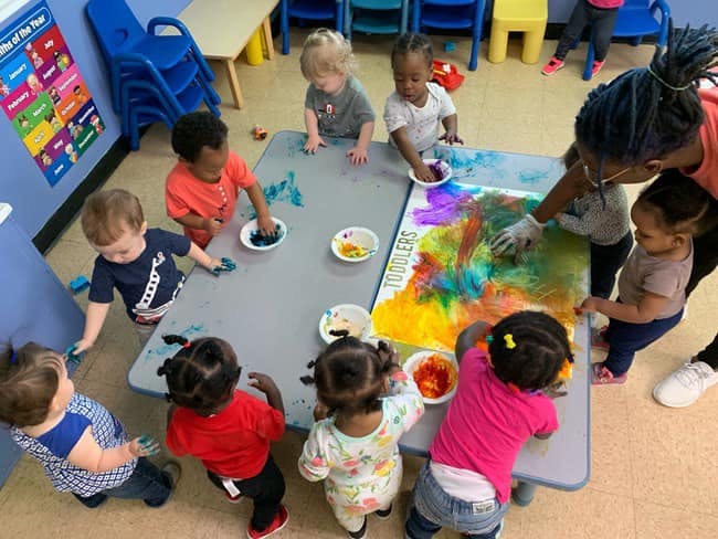 Adult and multiple children finger painting at a table in a classroom