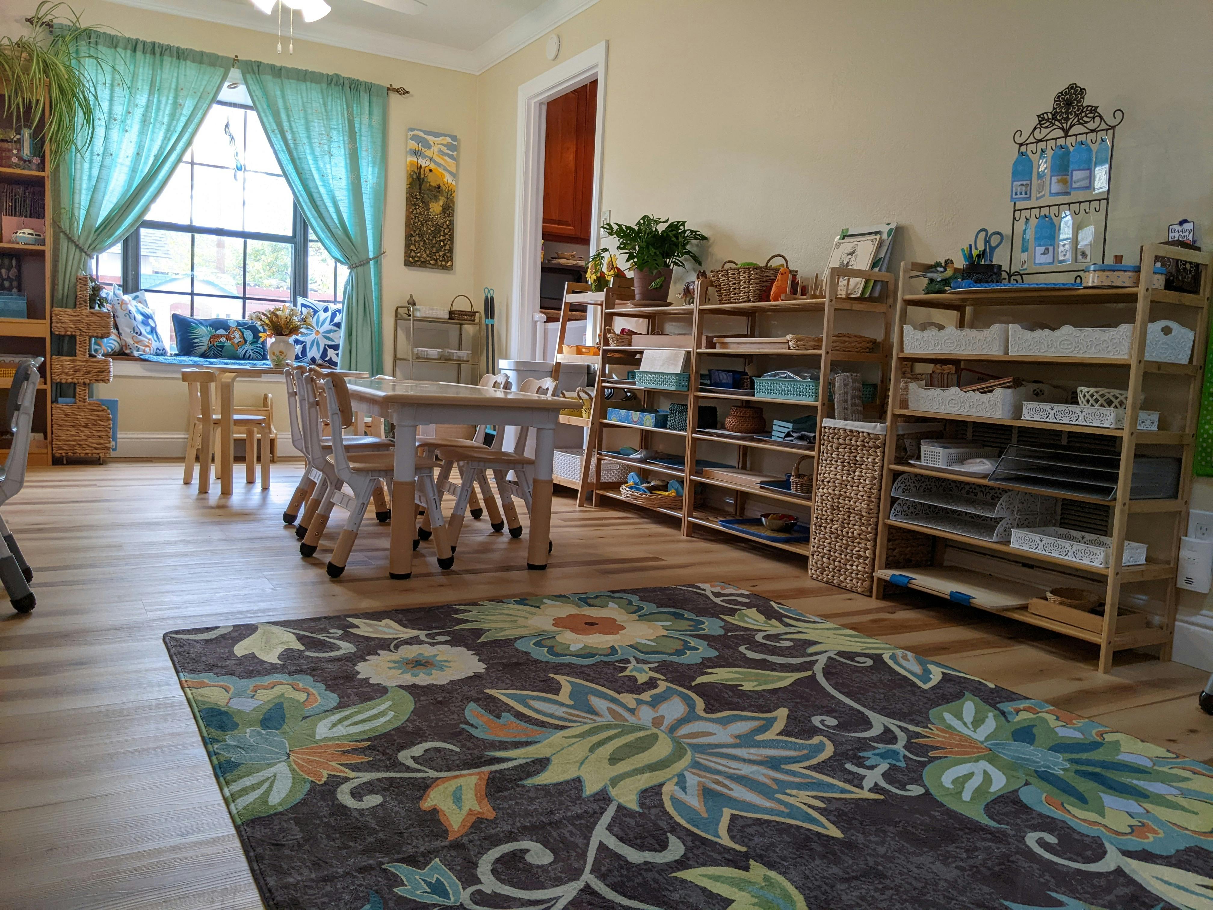 Classroom with small tables and chairs, shelves with learning materials, and a rug
