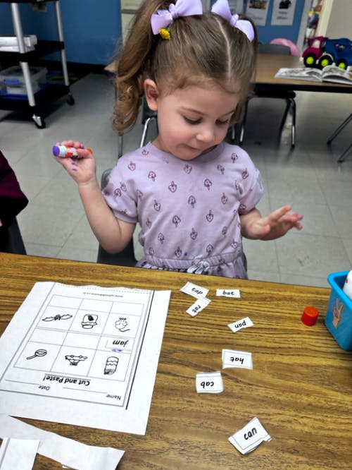 Child holding a glue stick, looking at a worksheet and word cutouts on a desk