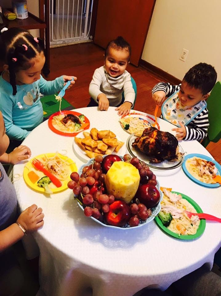 Three children eating at a table with plates of food and a fruit bowl