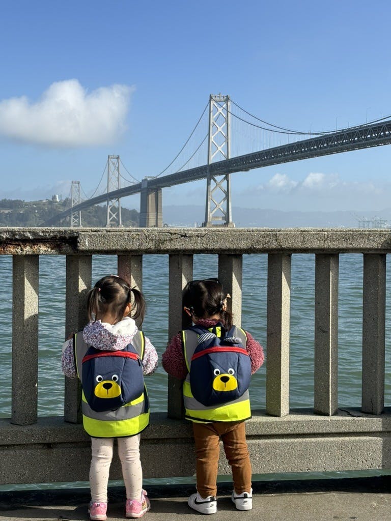 Two children in safety vests and backpacks looking at a bridge over water