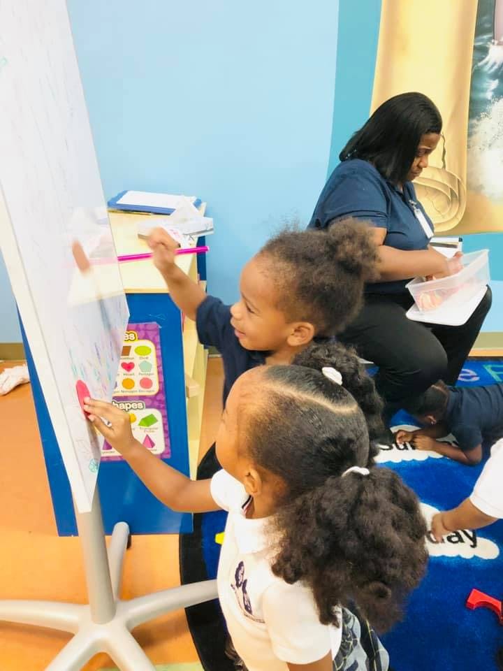 Two children drawing on a whiteboard in a classroom with an adult nearby