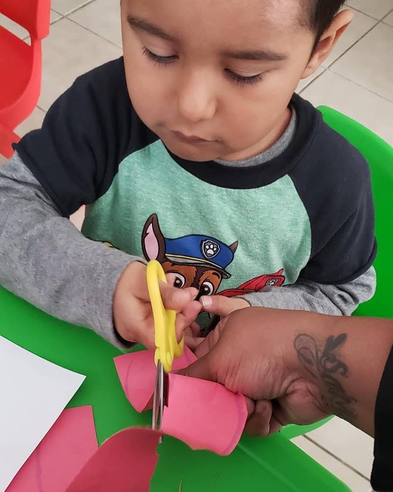 Child cutting pink paper with yellow scissors, assisted by an adult hand