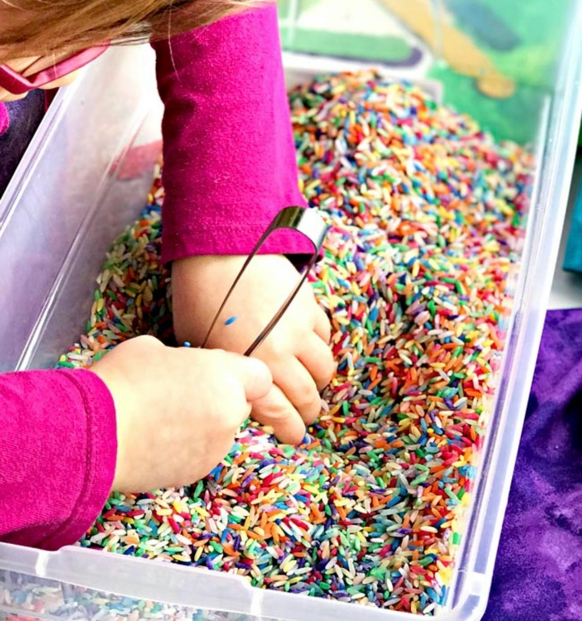 Child using tweezers to pick up grains of colorful rice from a plastic bin