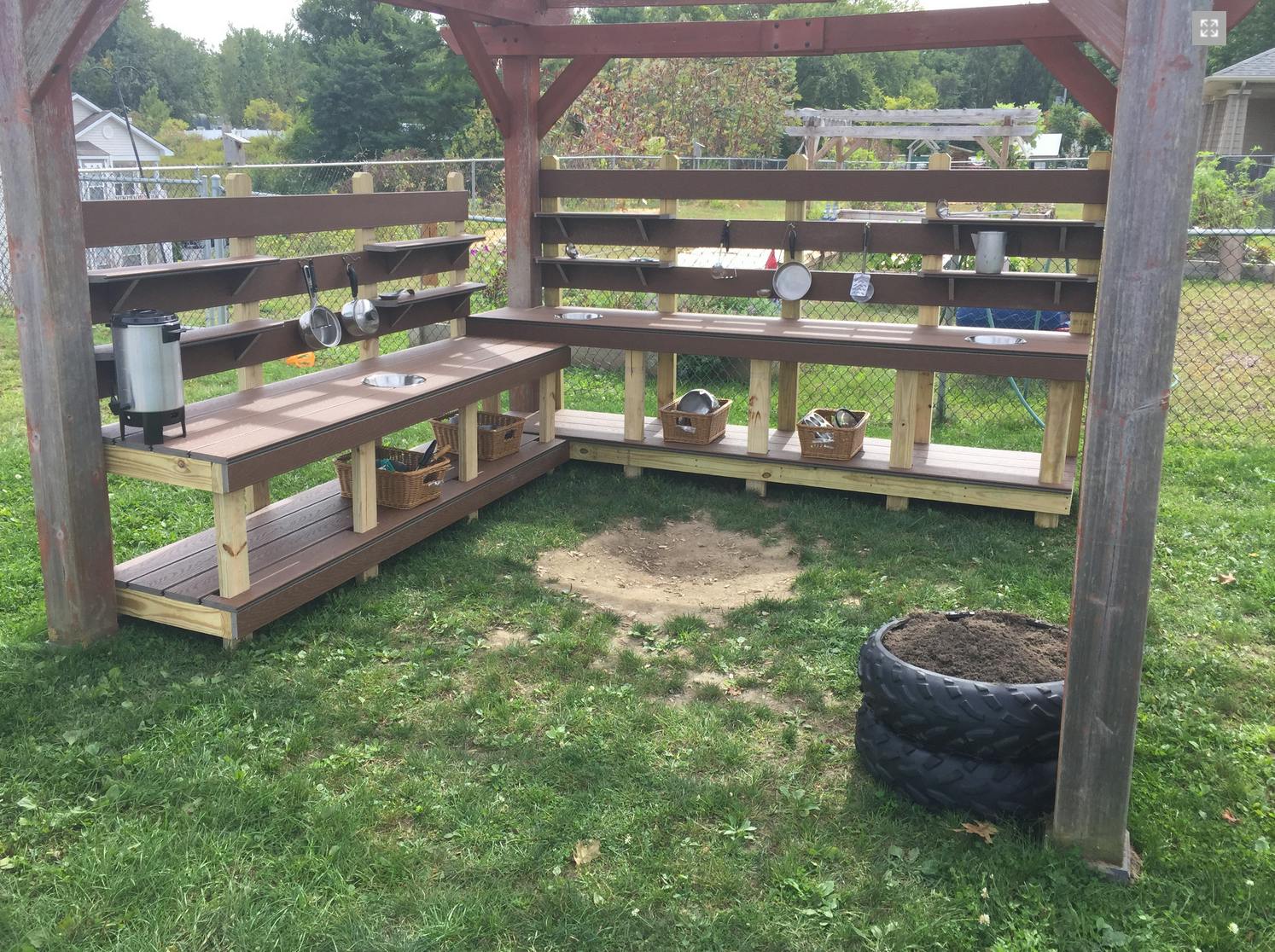 Outdoor mud kitchen with wooden benches and shelves, sand pile, and tire planter