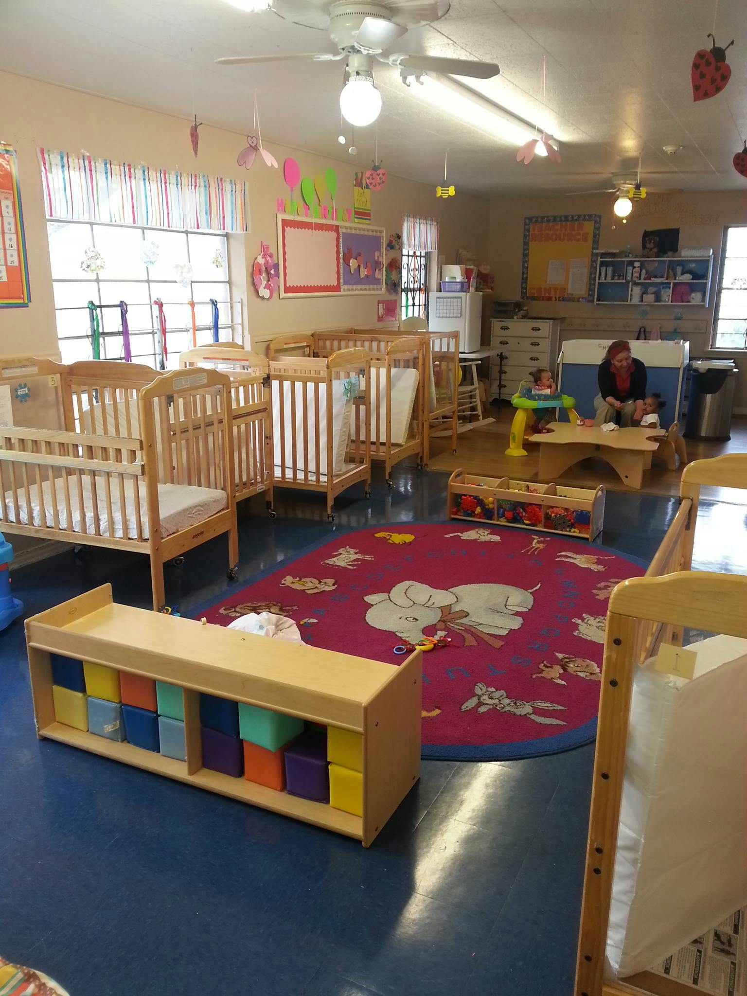 Daycare room with multiple cribs, a colorful rug, and a person with a child