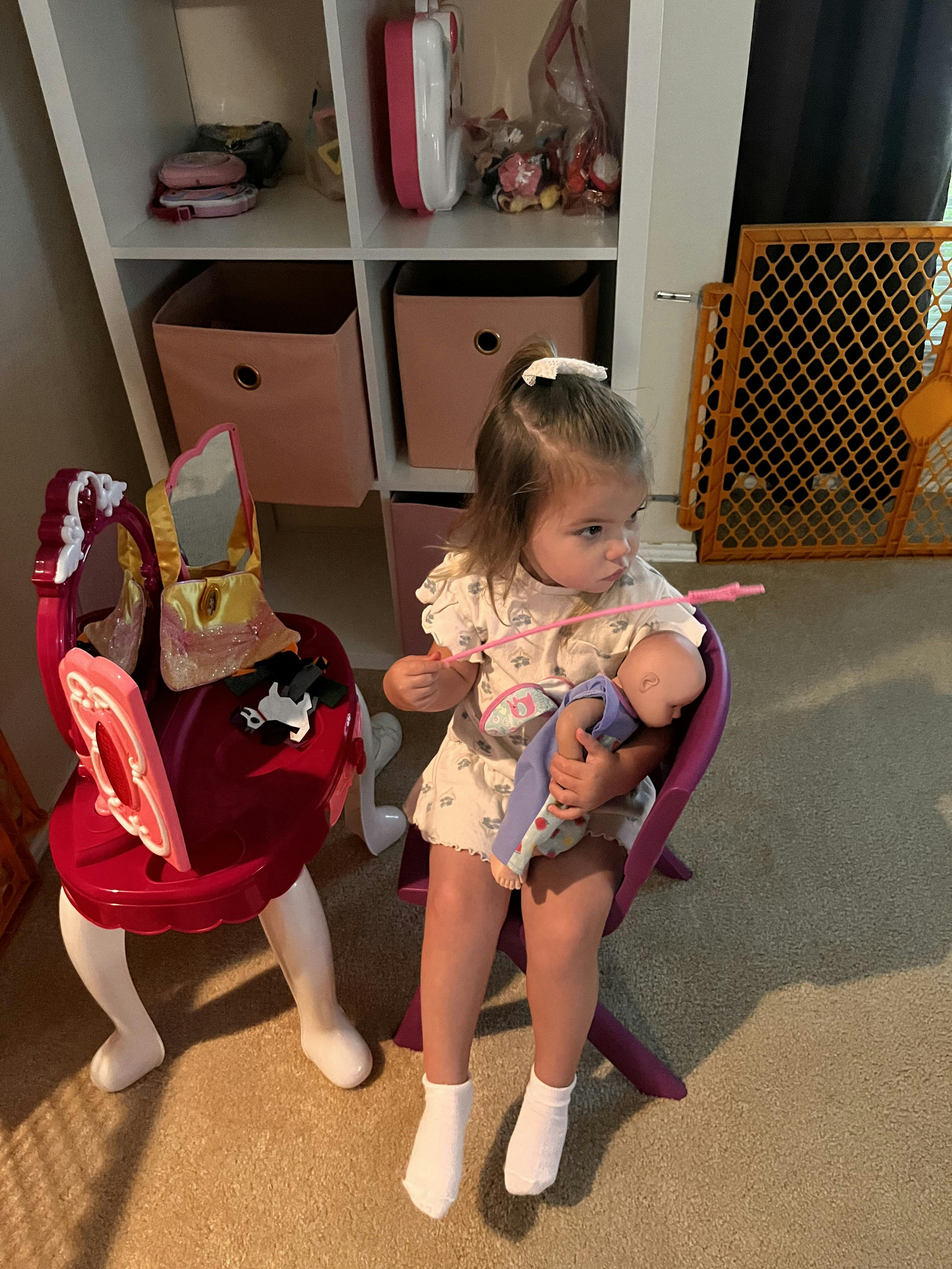 Child sitting on a chair holding a doll and a toy wand