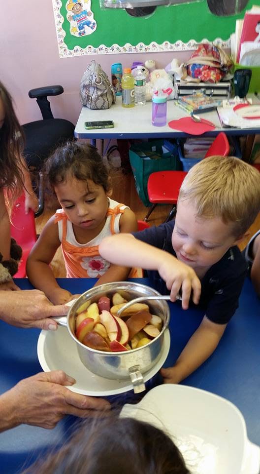 Two children at a table with an adult holding a pot of sliced apples