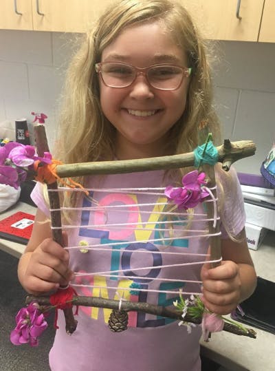 Child holding a craft made from sticks, string, and flowers