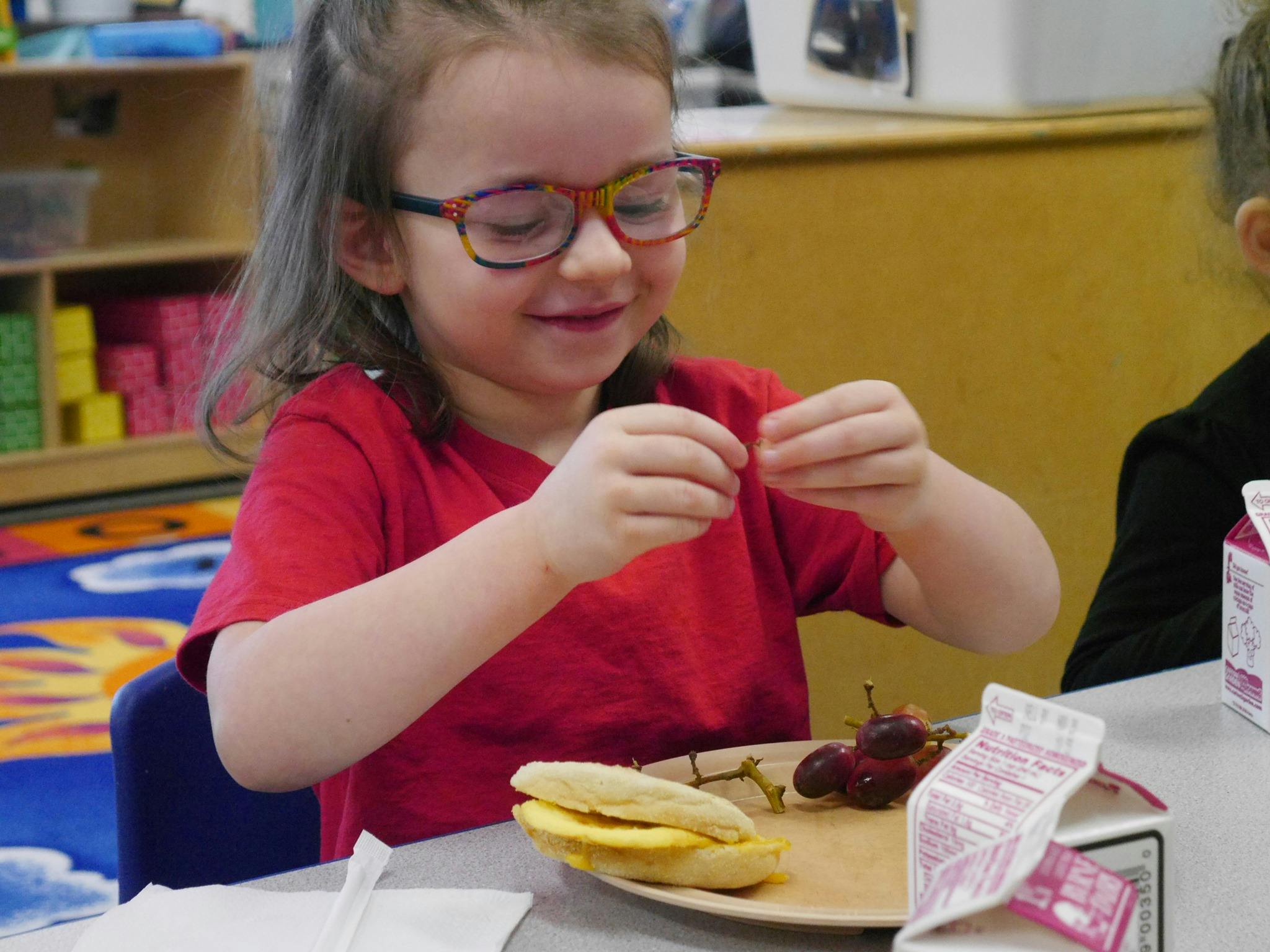 Child in glasses holding food over a plate with a sandwich and grapes