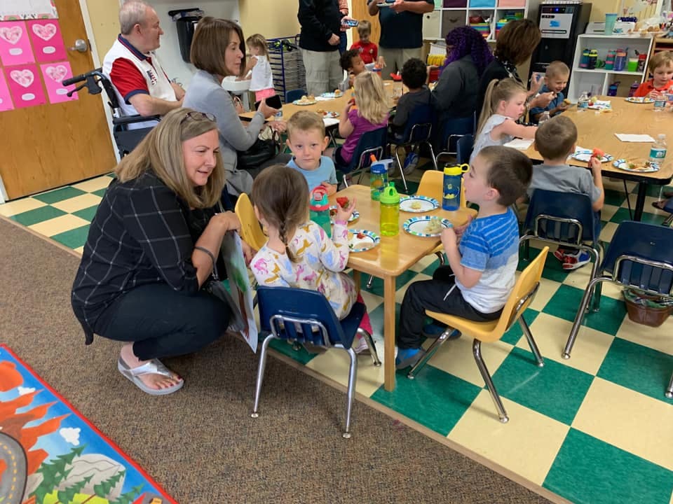 Adult interacting with children seated at small tables in a classroom