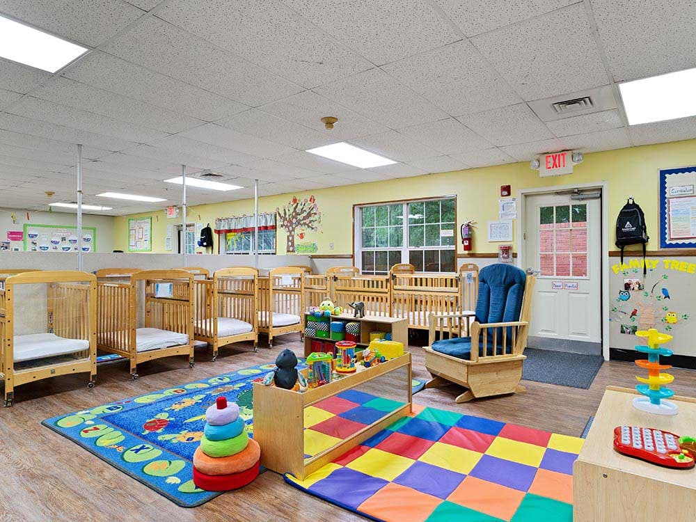 Cribs and colorful playmats in a daycare classroom with yellow walls