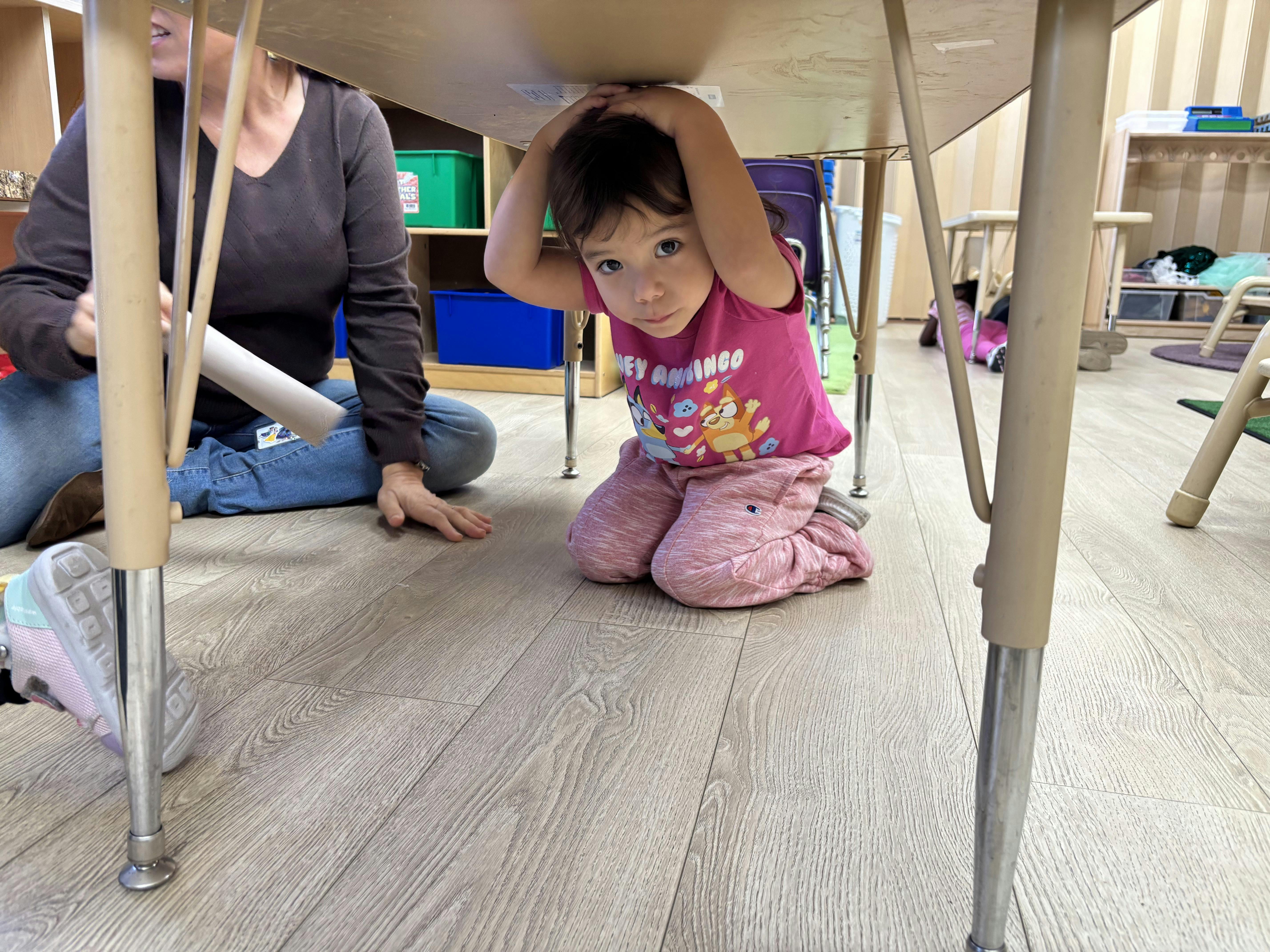 Child kneeling under a table with hands on head, person sitting nearby