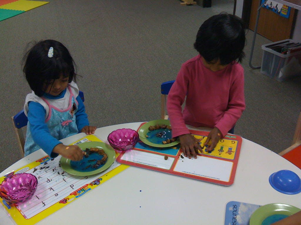 Two children at a table, one painting a plate, the other playing with a toy