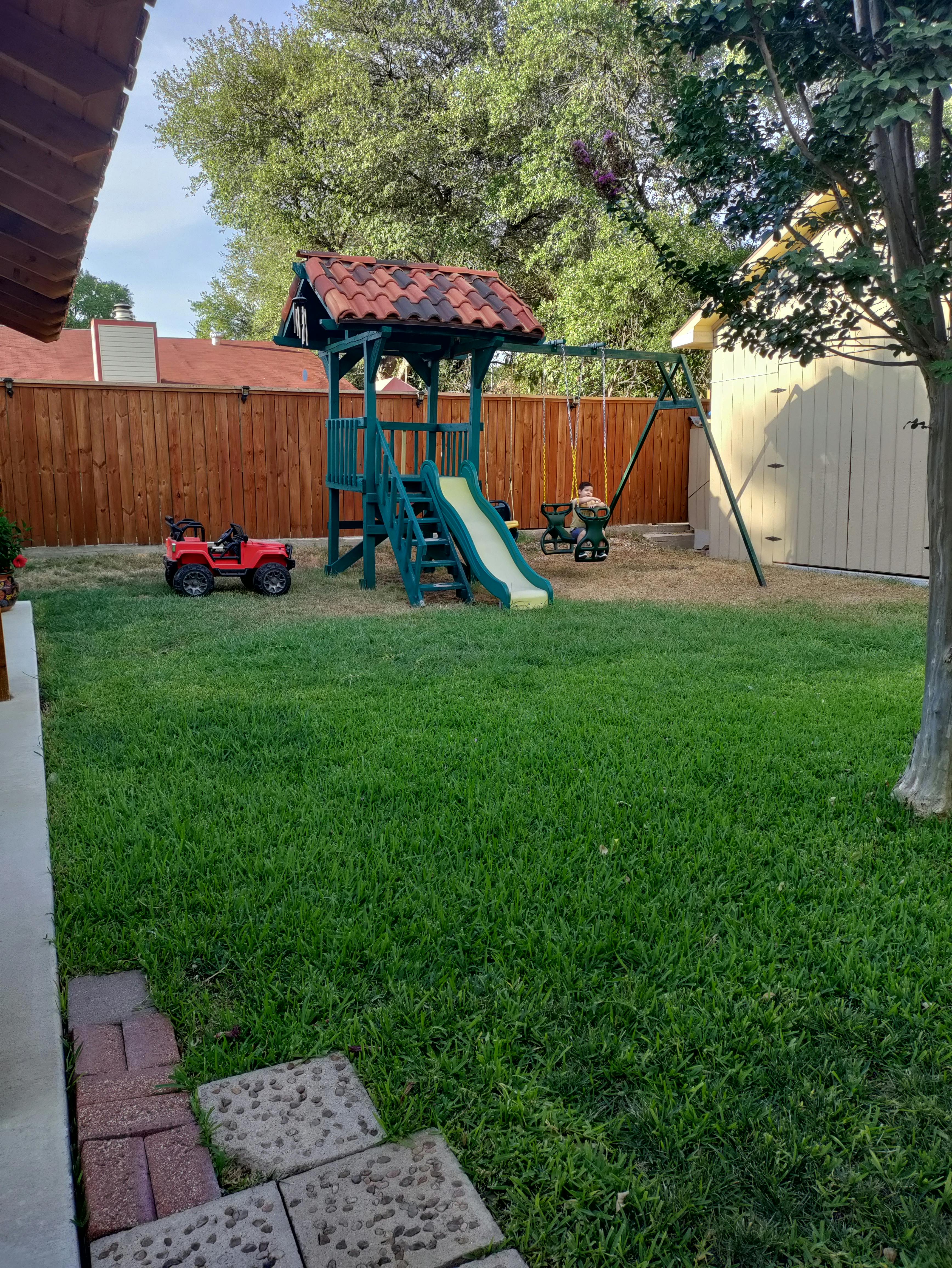 Backyard with a wooden playset, slide, swings, and a red toy car