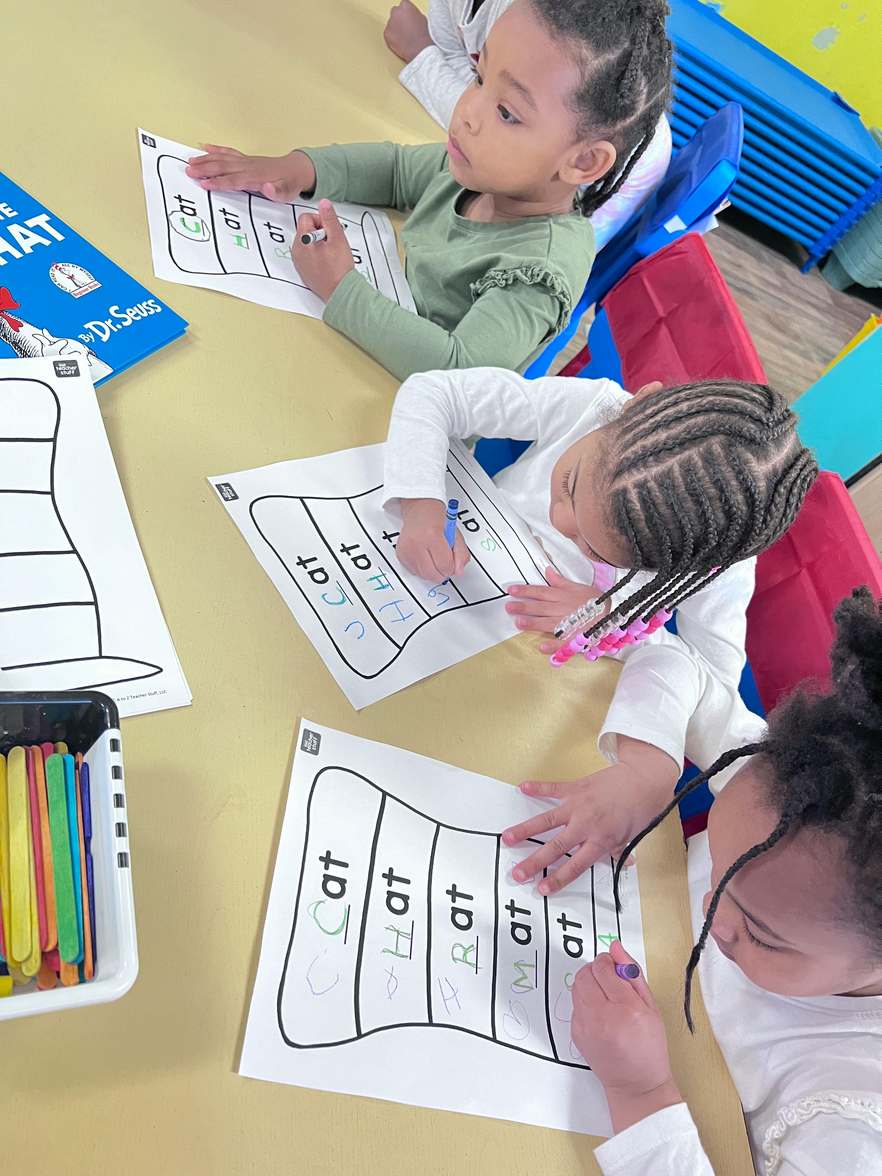 Three children tracing words on hat-shaped worksheets in a classroom