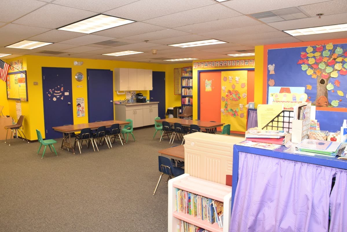 Classroom with small tables and chairs, yellow walls, and a blue bulletin board