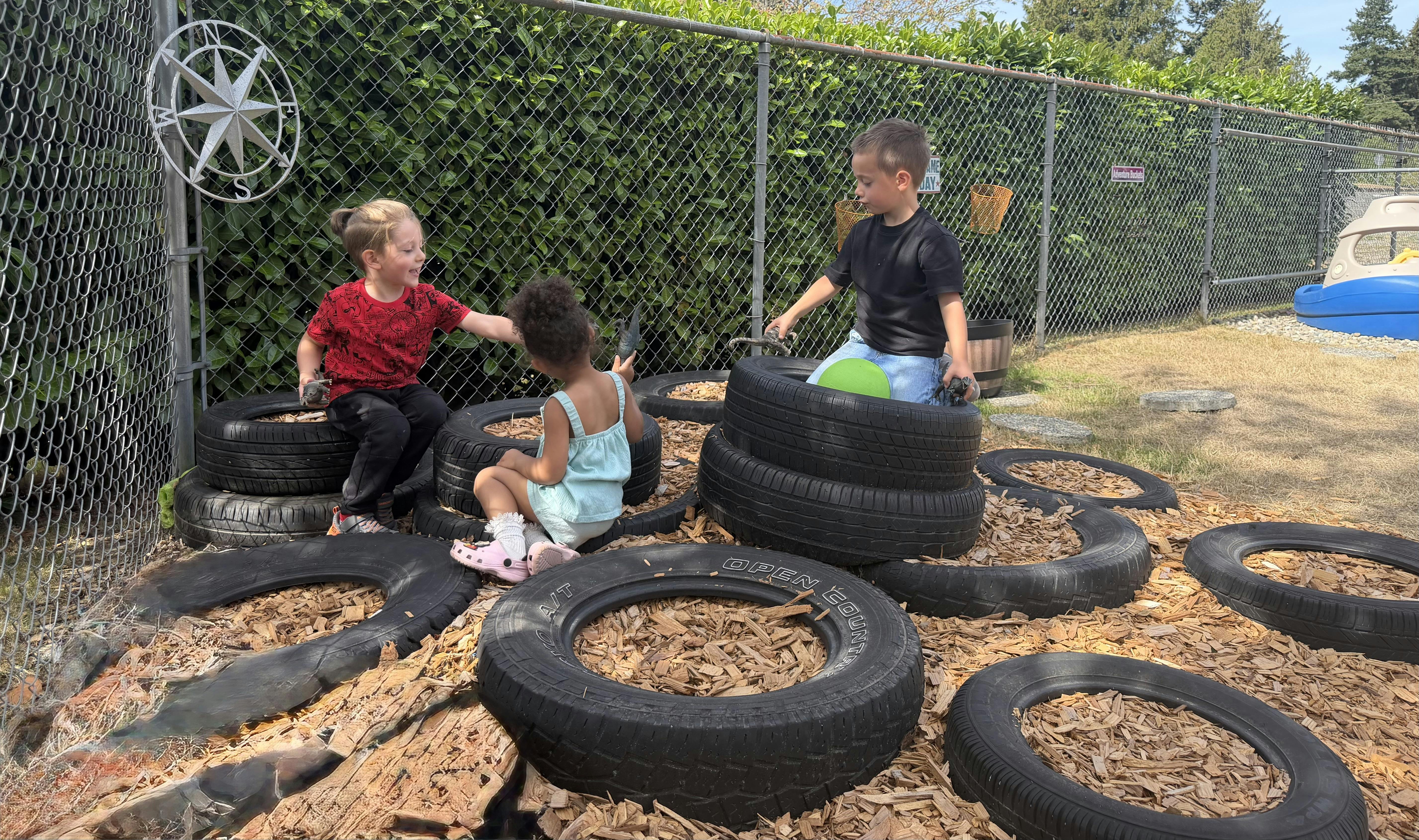 Three children playing with toy dinosaurs on and around tires in a fenced area