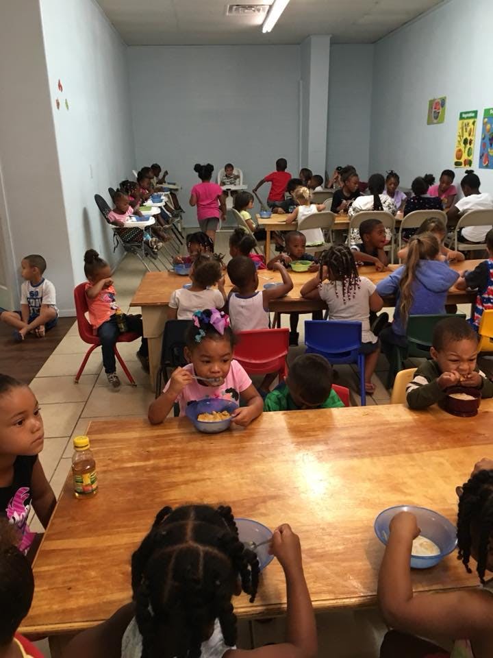 Many children sitting at tables eating in a room, some eating from bowls