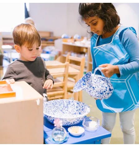 Child in apron pouring from a pitcher into a bowl, another child watching