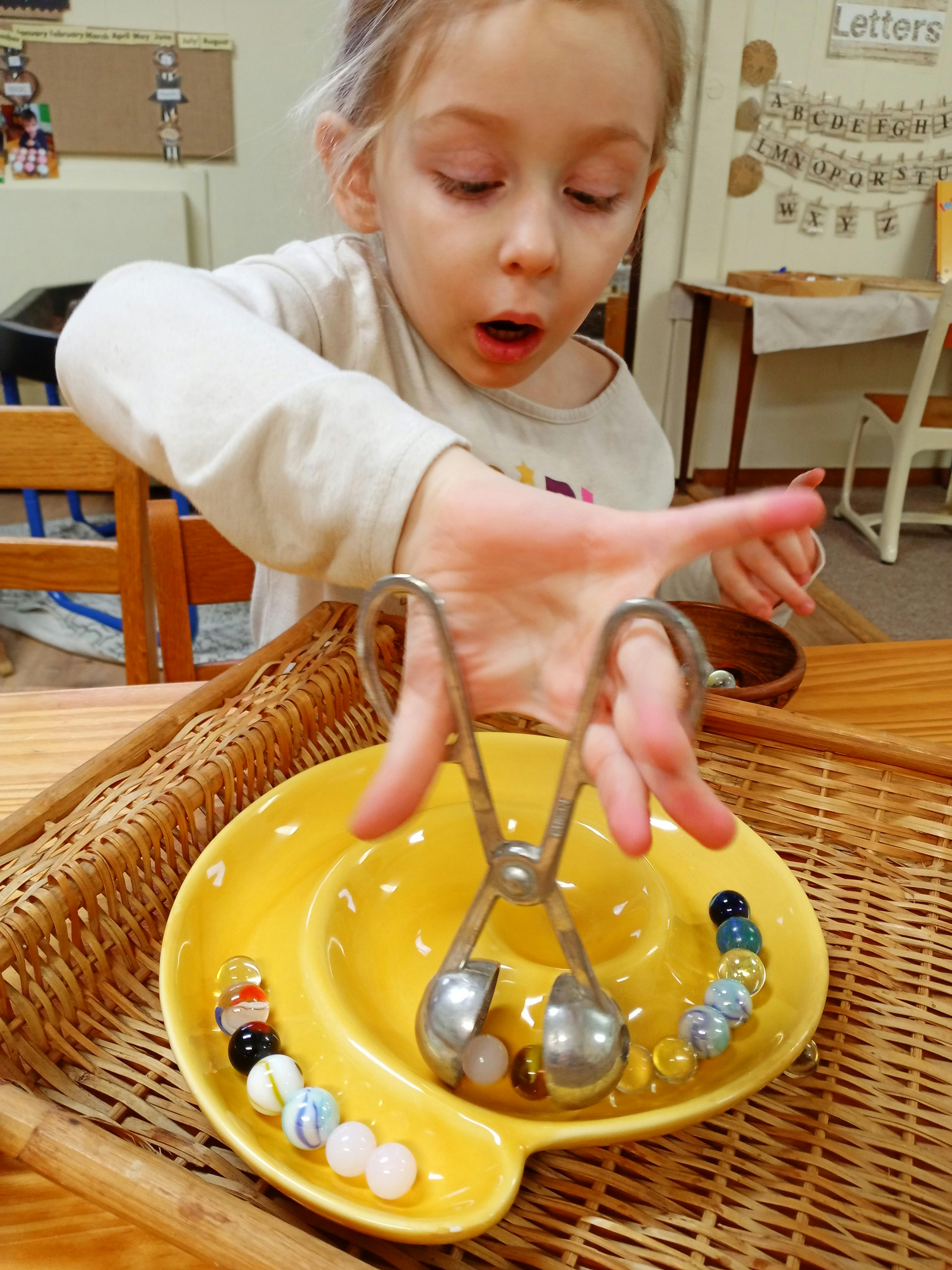 Child using tongs to pick up marbles from a yellow dish