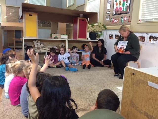 Adult reading a book to children sitting on the floor, some with hands raised