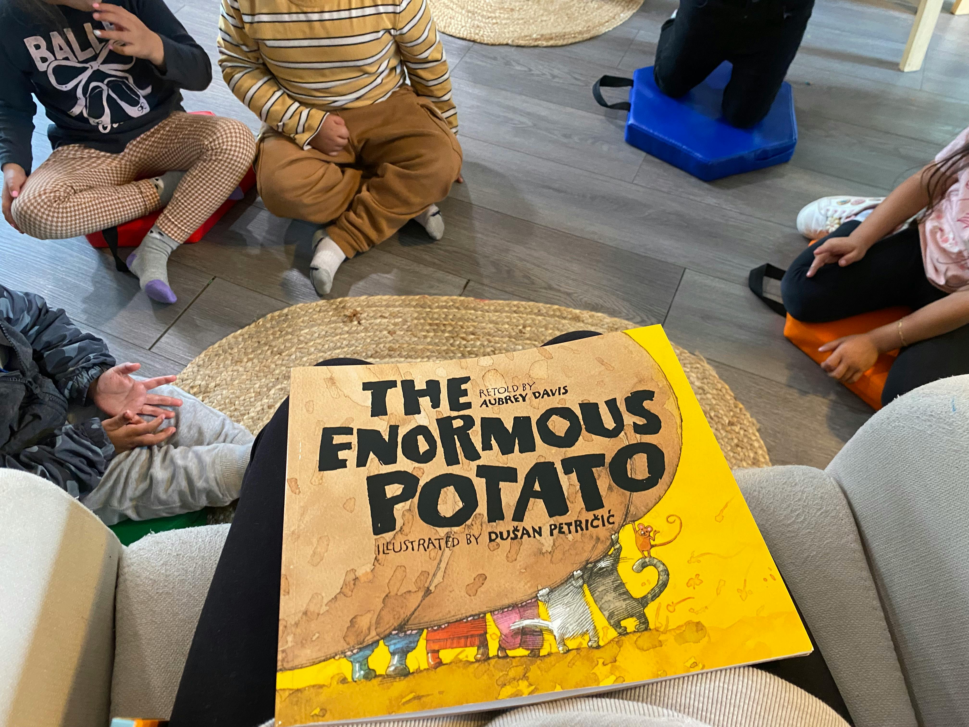 Children sitting on floor around a book titled The Enormous Potato