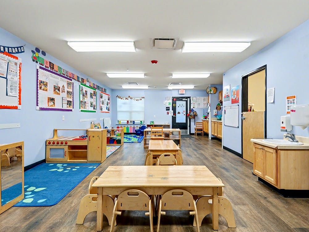 Classroom with small wooden tables and chairs, play kitchen, and bulletin boards