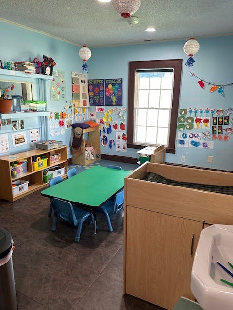 Classroom with a green table and blue chairs, shelves with toys, and decorated walls