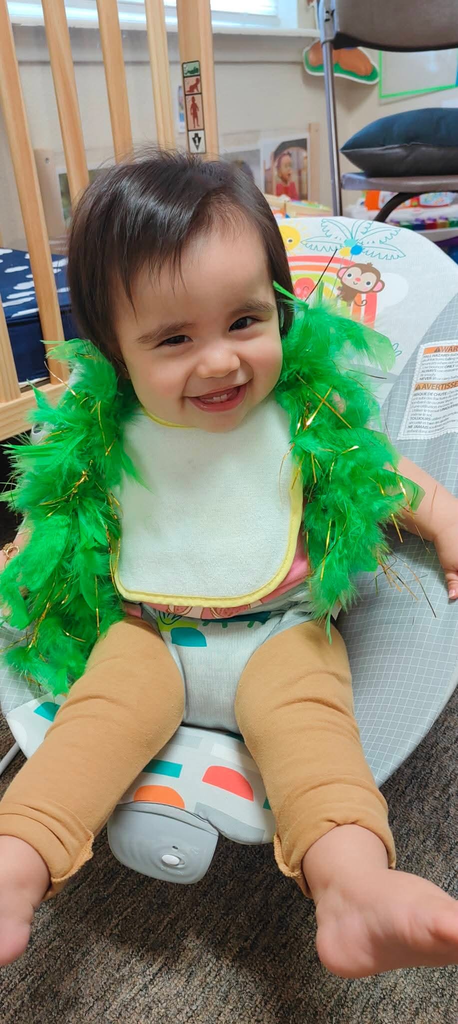 Child wearing a green feather boa and a bib, sitting in a baby bouncer