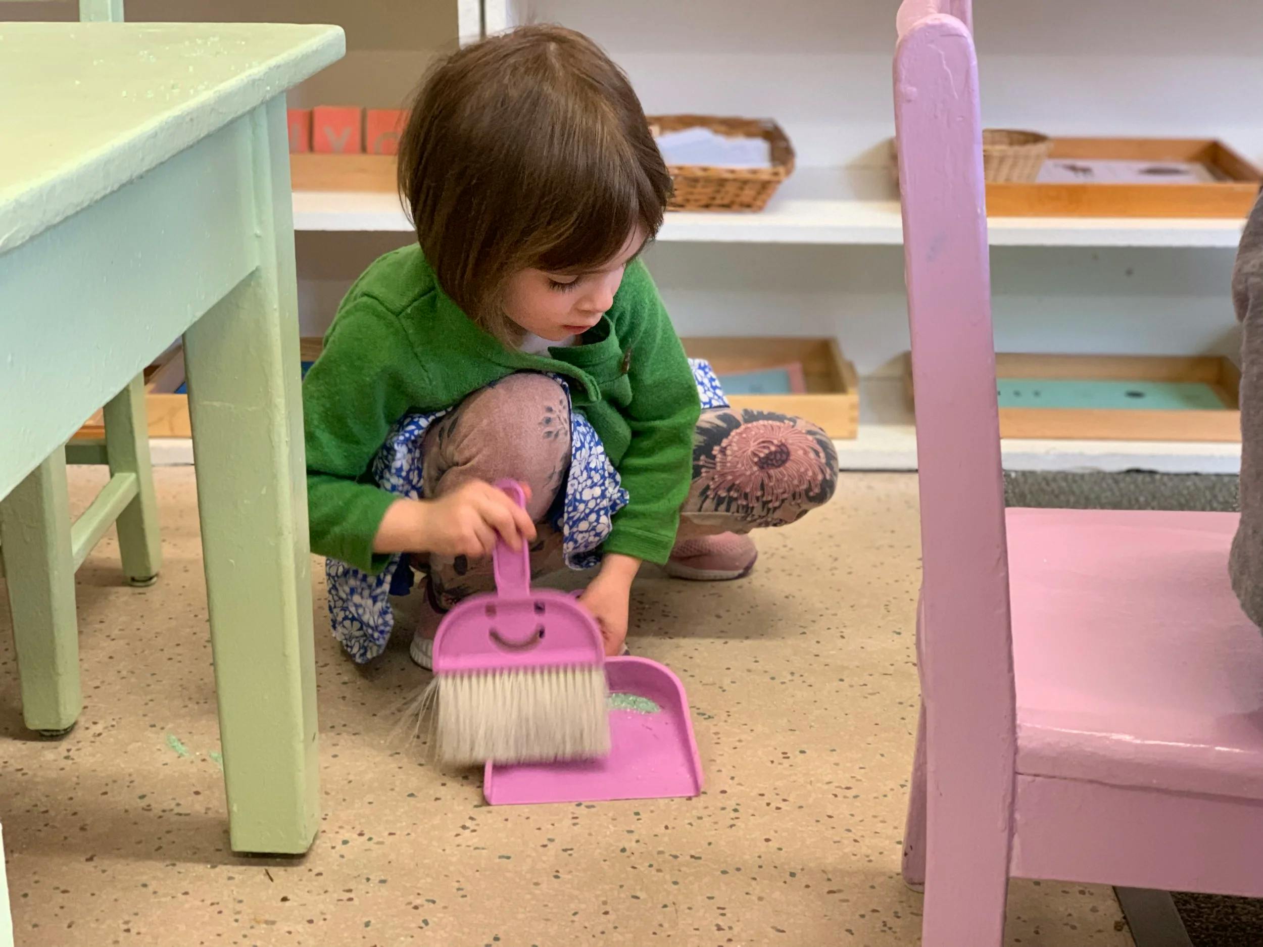 Child sweeping small green beads with a pink toy broom and dustpan
