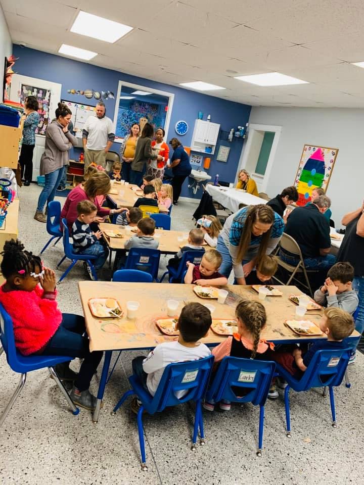 Adults and children gathered in a classroom, some seated at tables with plates