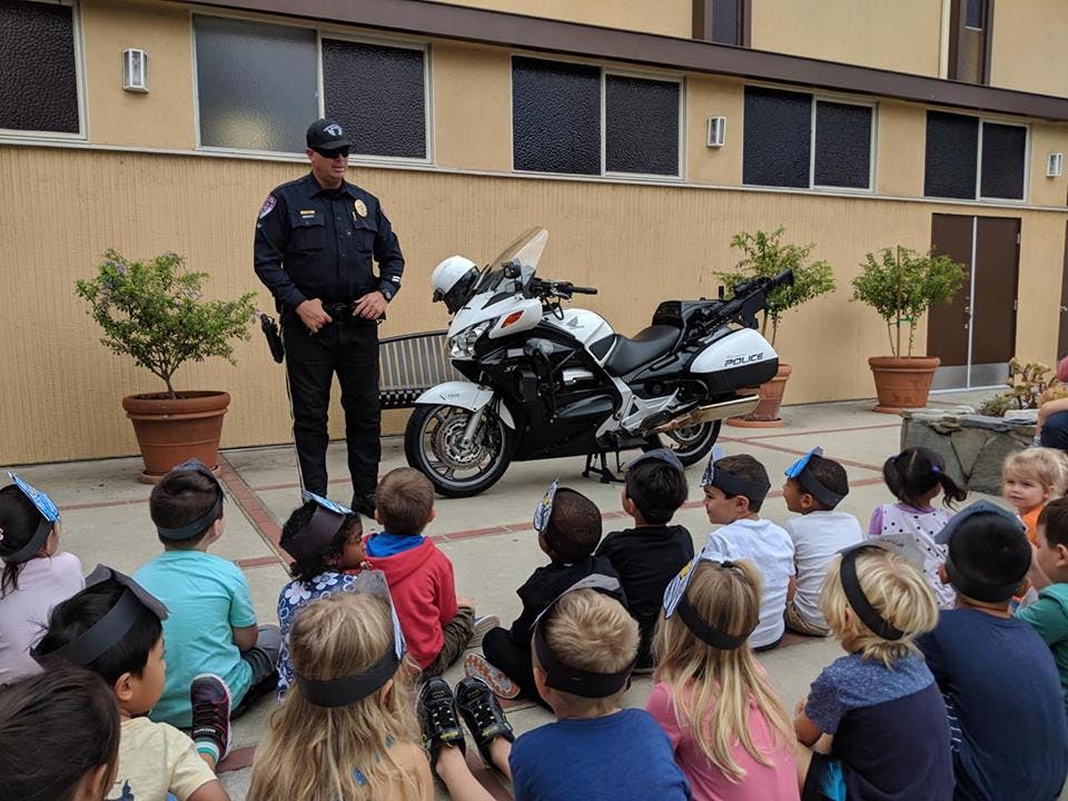 Person in uniform standing next to a motorcycle, speaking to children sitting on the ground