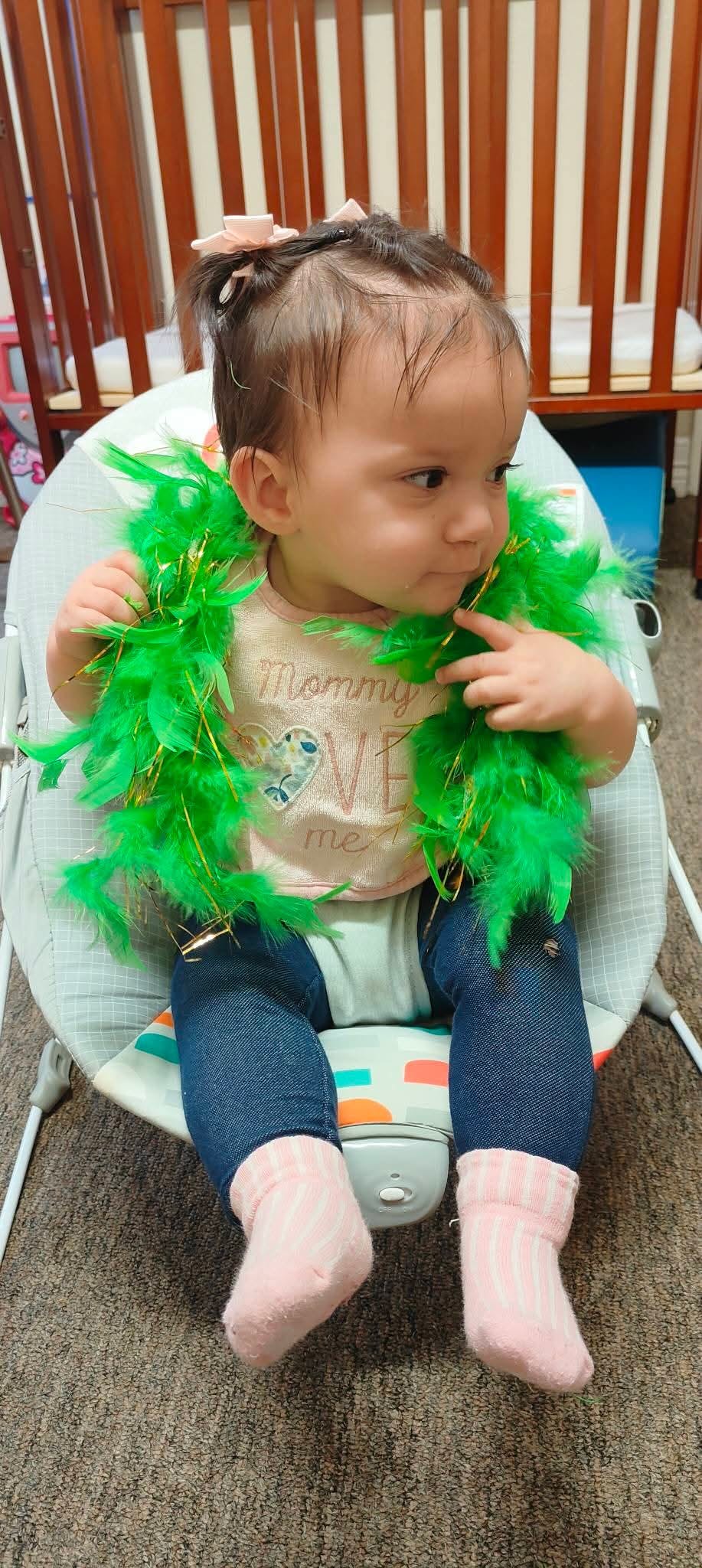 Child sitting in a bouncer wearing a green feather boa