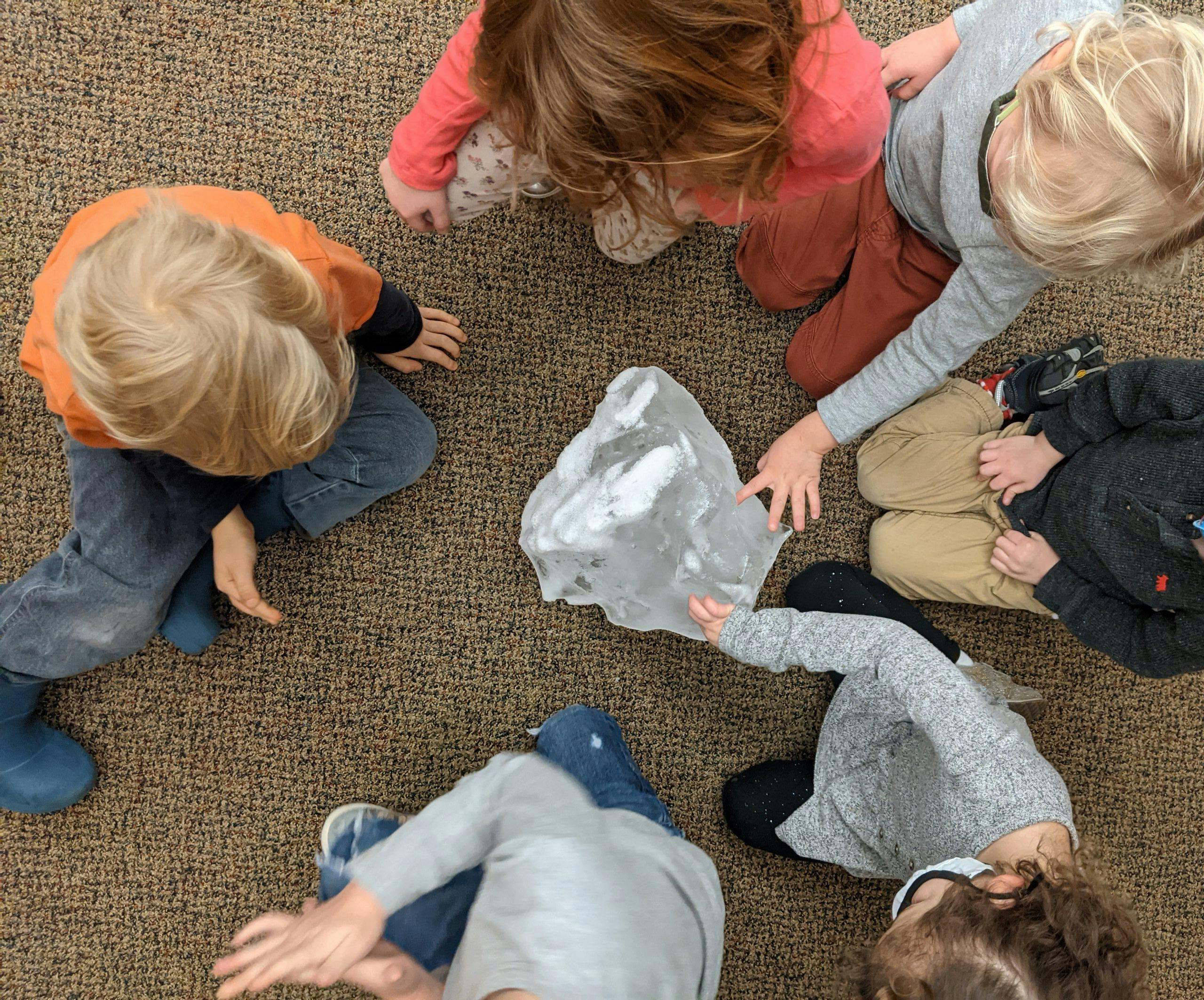 Several children gathered around a large block of ice on a carpeted floor
