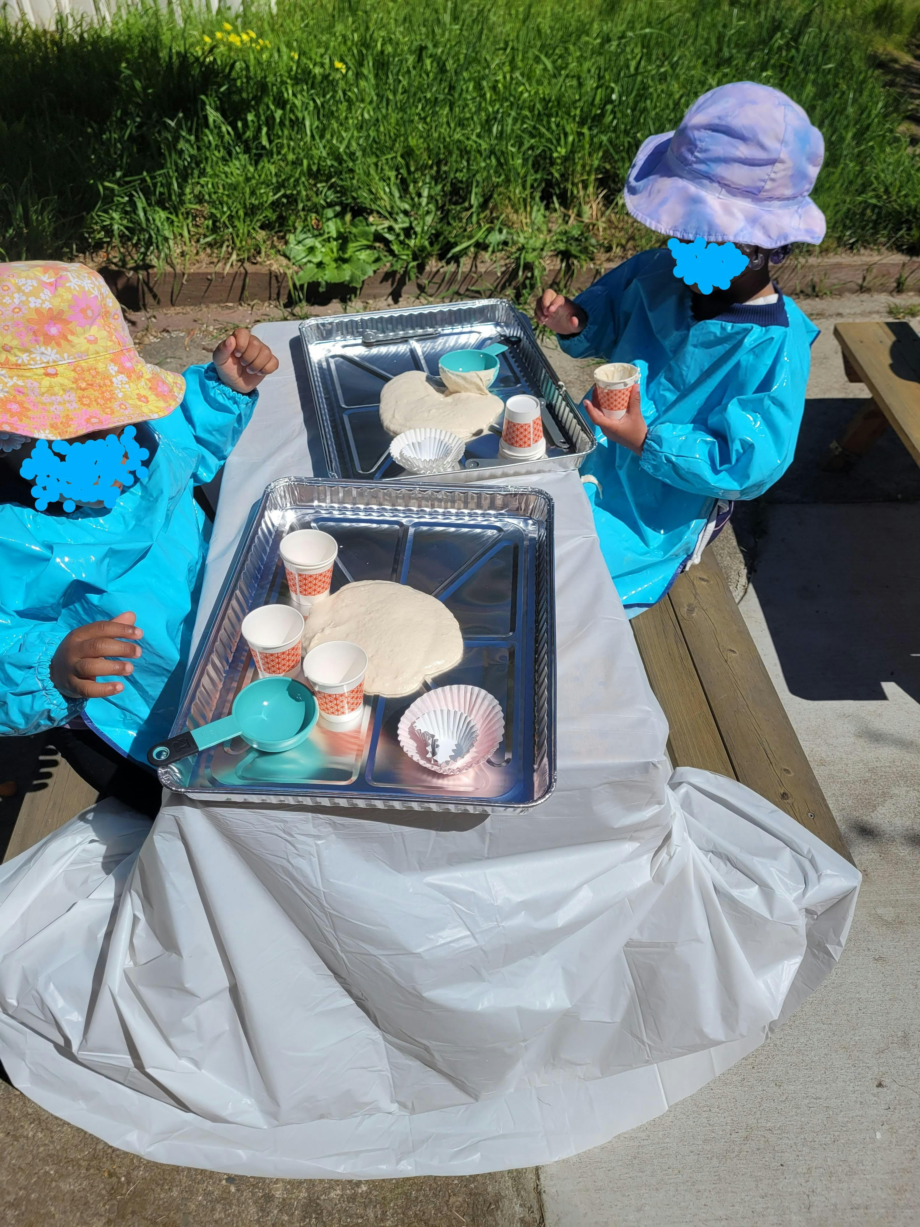 Two children wearing smocks and sun hats playing with dough on outdoor trays