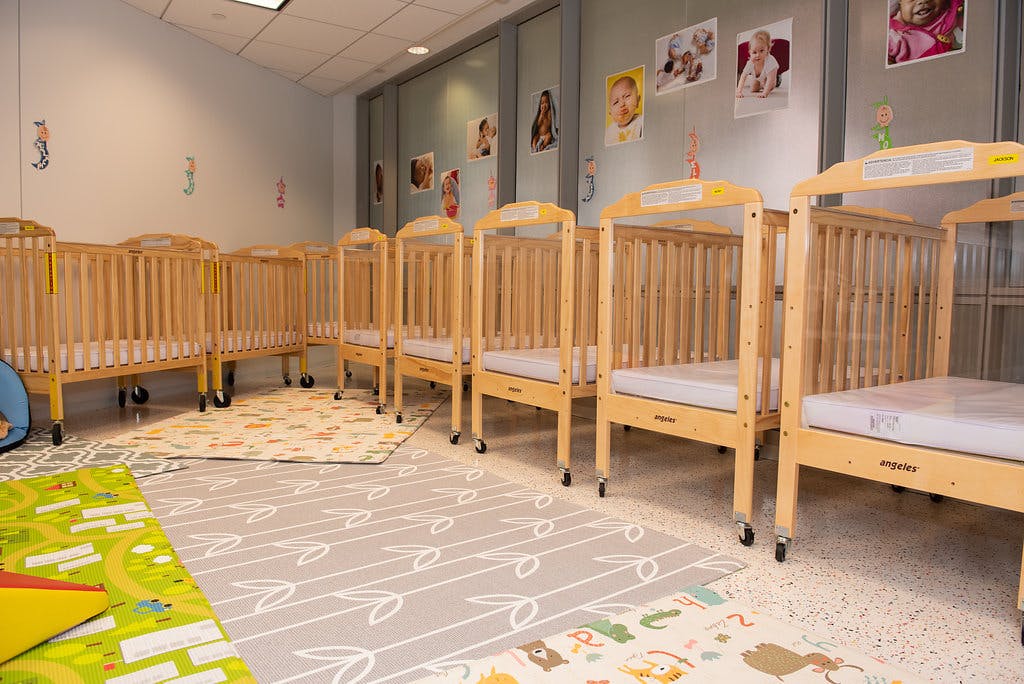 Many empty cribs lined up in a bright room with colorful play mats on the floor