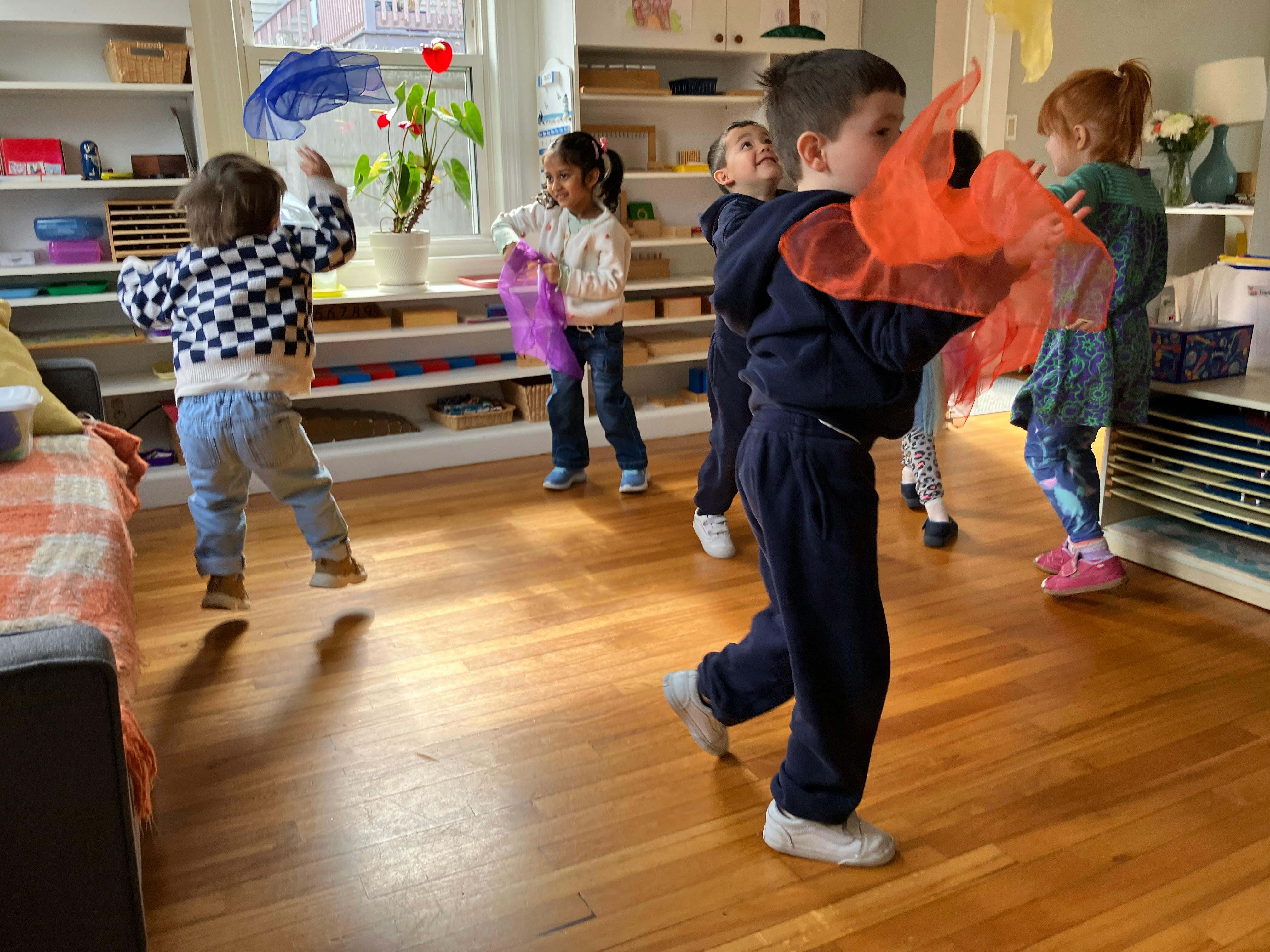 Group of children playing in a classroom, some holding colorful sheer fabric