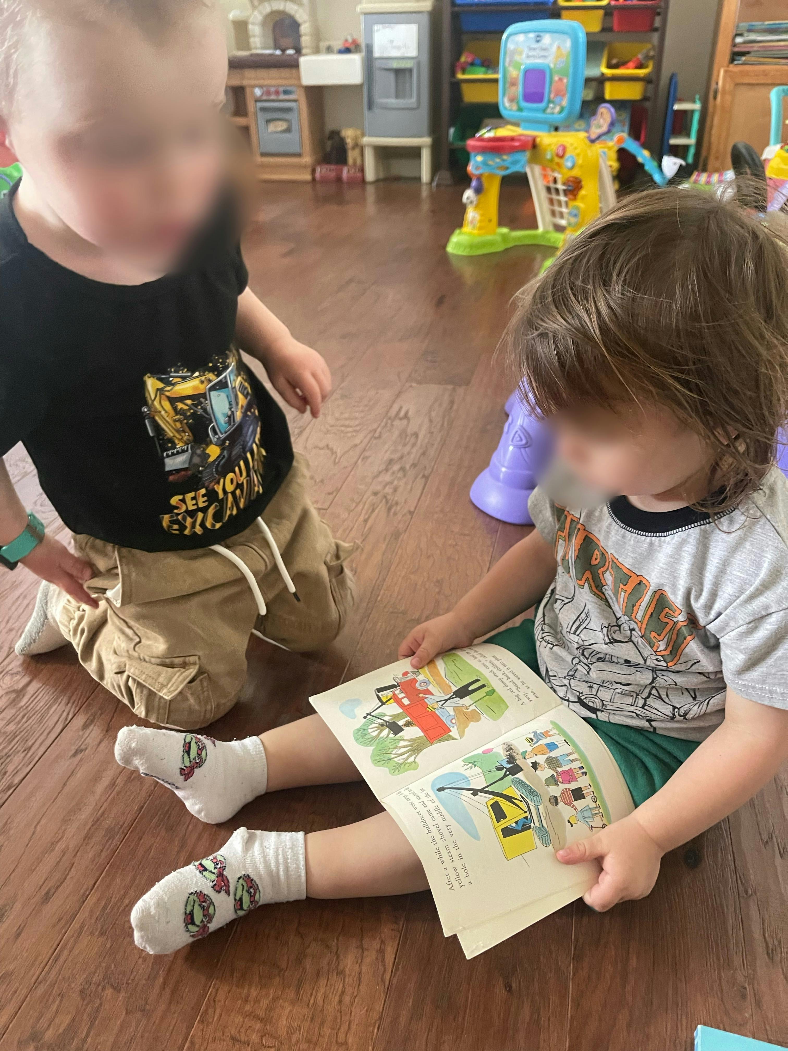 Child sitting on floor reading a book, another child kneeling nearby