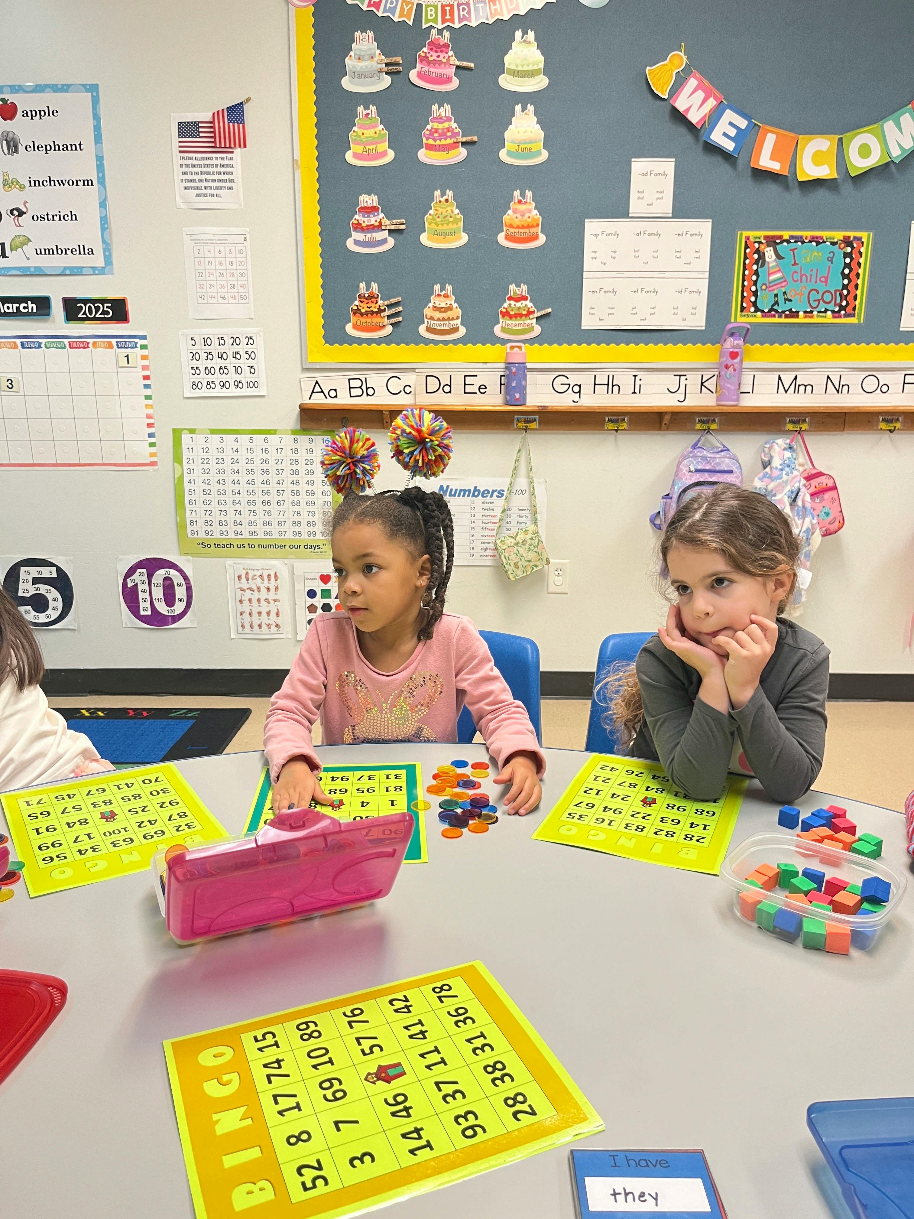 Two children at a table with bingo cards and colorful game pieces in a classroom