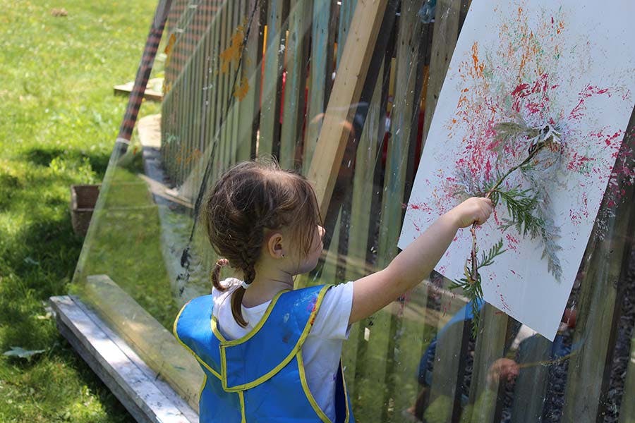 Child painting with a plant branch on an easel outdoors