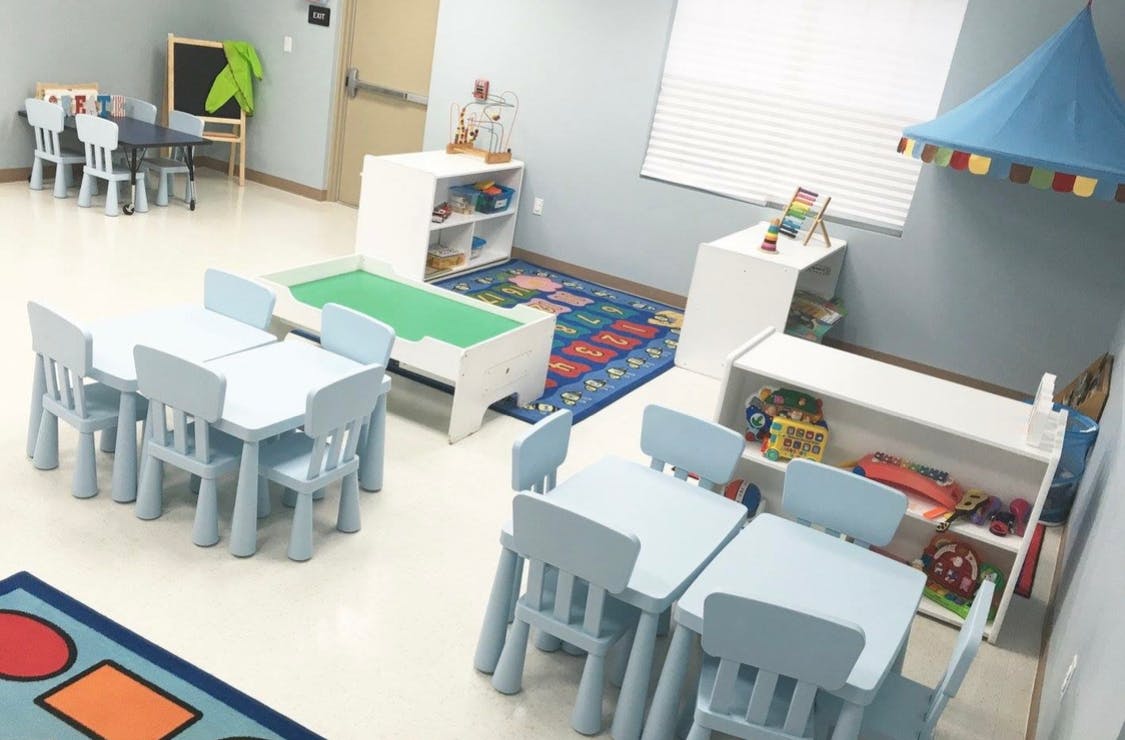 Empty classroom with small tables and chairs, play rug, and shelves