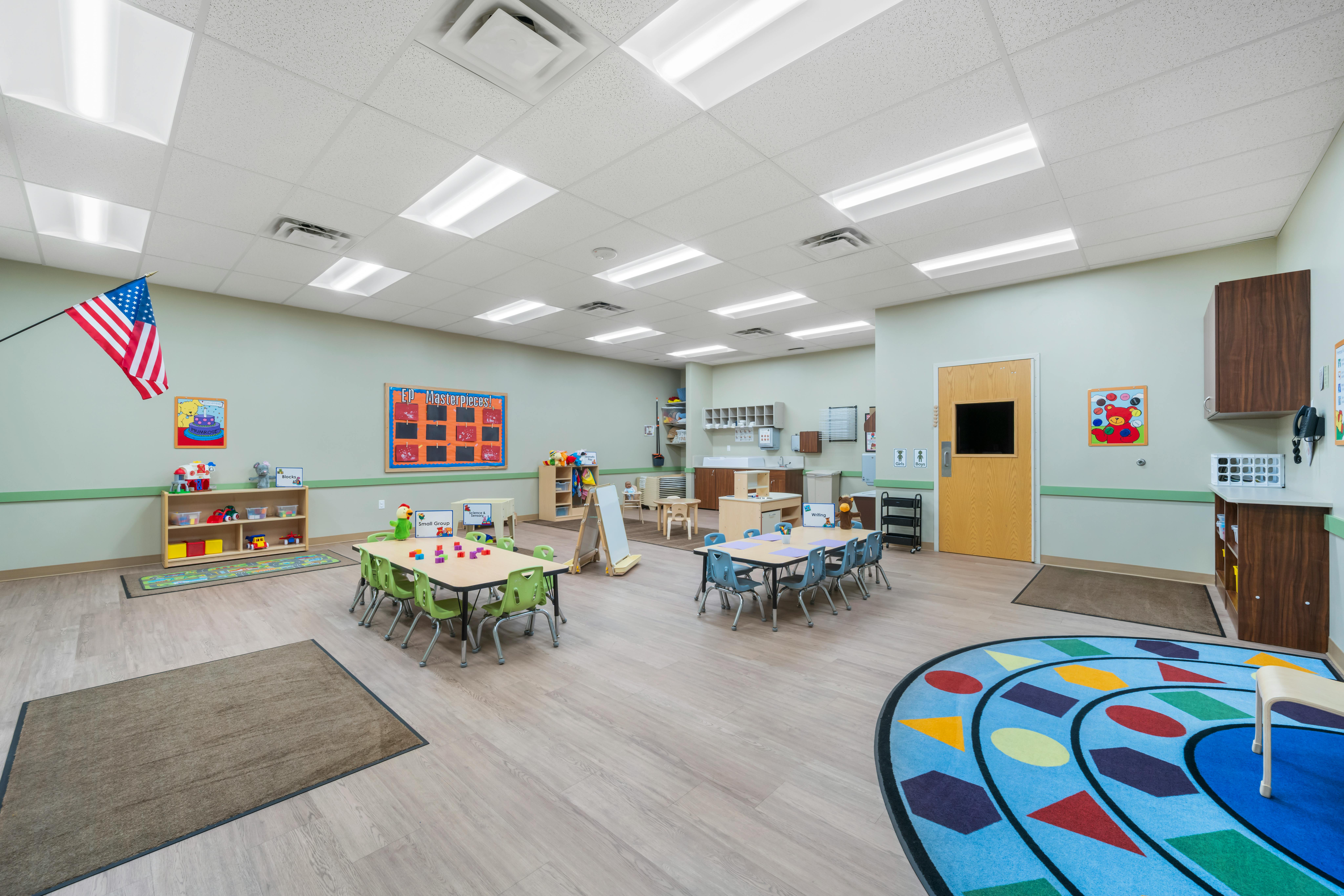 Classroom with small tables and chairs, shelves with toys, and an American flag