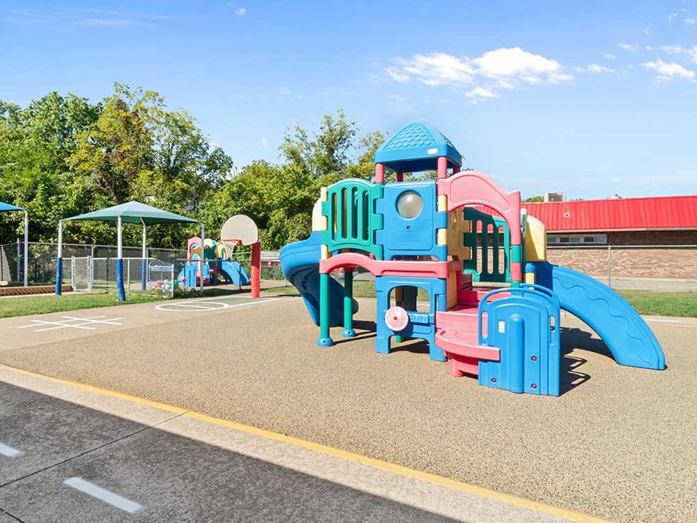 Colorful playground structure on a tan soft surface with trees and a building in the background