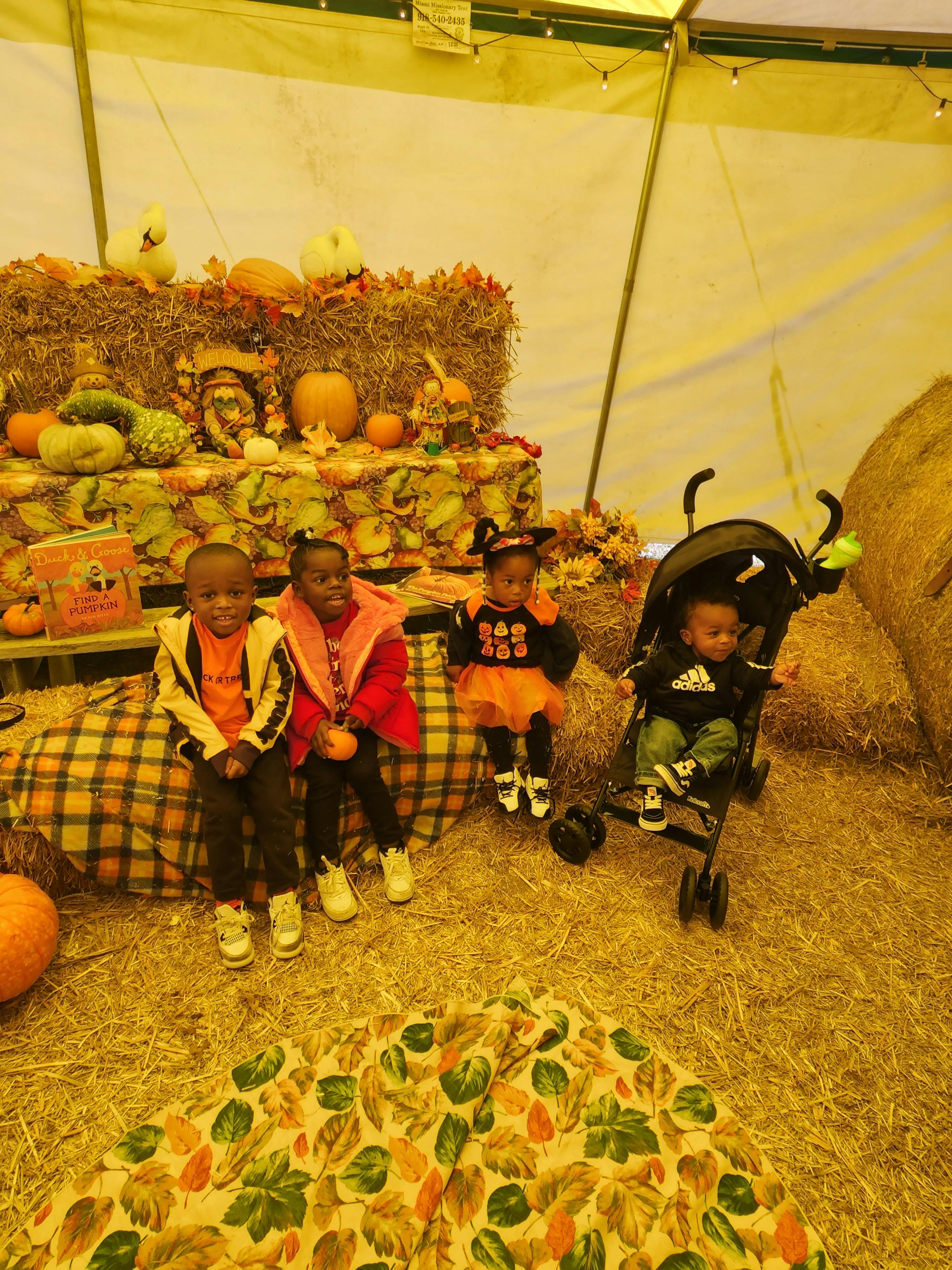 Three children and a baby in a stroller at a fall display with hay bales and pumpkins