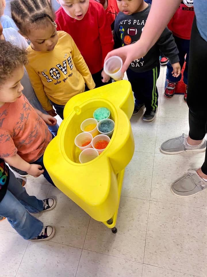 Children gathered around a yellow container with cups of colorful materials
