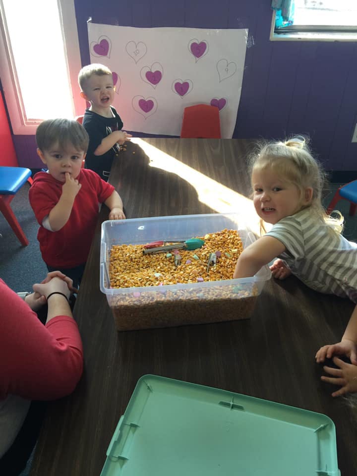 Three children playing with a sensory bin filled with corn kernels