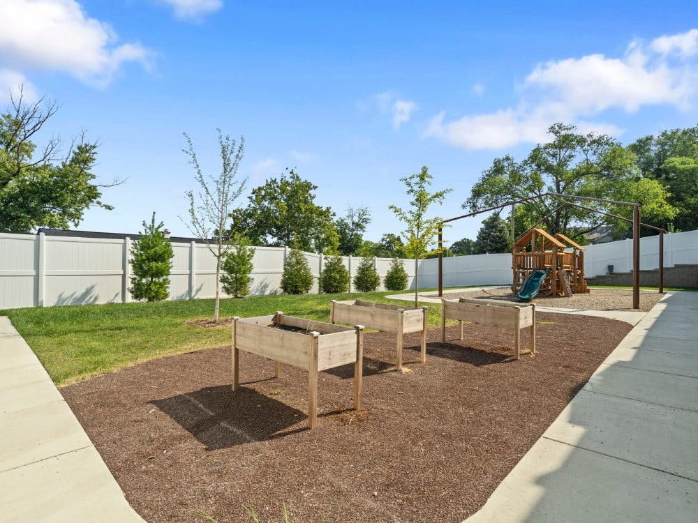 Raised garden beds on wood chips and a playground behind a white fence