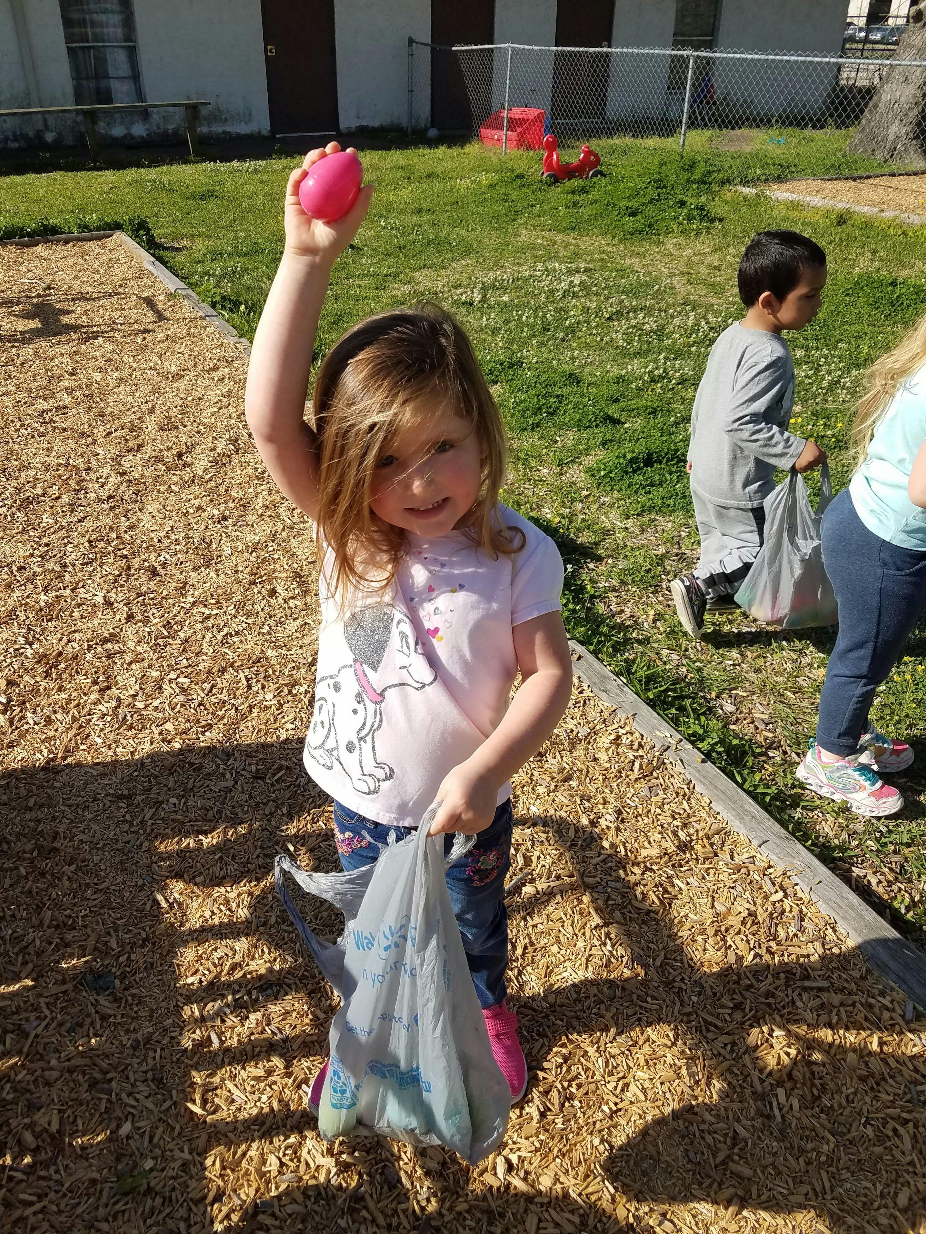 Child holding up a pink egg and carrying a bag outdoors